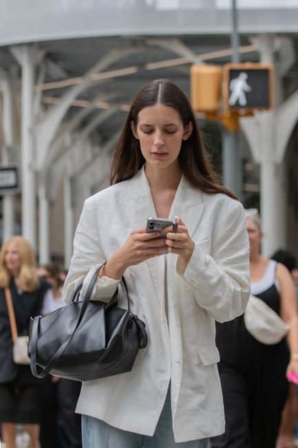 A woman with long dark hair, dressed in a light beige blazer and blue jeans, is holding a smartphone with both hands and looking at the screen. She carries a black leather handbag on her left shoulder