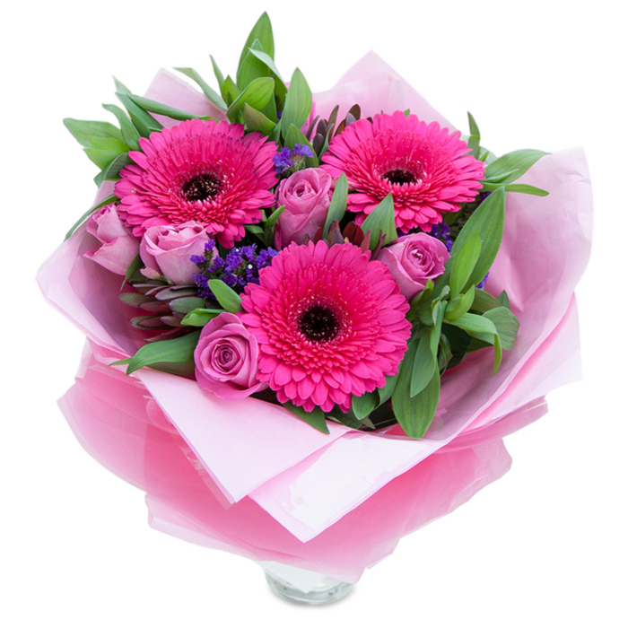 I see this bouquet as a small homage to garden charm, where playful colour meets considered structure. Three wide-faced Gerbera daisies in saturated fuchsia open confidently at the heart of the design, their many petals radiating around deep, almost inky centres. Surrounding them, lavender-tinged roses in a dusty rose hue curl inward with the innocence of early blooms, their layered petals adding softness and romantic rhythm. Little sprays of purple statice tuck into the spaces between larger flowers, lending a textured, papery contrast and a hint of wildflower spontaneity. The foliage is generous and green - broad salal leaves and the cool grey-green of eucalyptus - which gives the bouquet weight and a fresh, slightly dewy aroma. The blooms are wrapped in an inner sheet of pale pink matte tissue and enveloped by a translucent fuchsia-pink cellophane that catches and diffuses light, creating a faint shimmer over the petals. Where the wrapping folds around the stems you can sense the careful hands that arranged it, and the top rim of a clear glass vase is visible at the base, implying that these flowers are intended to be enjoyed immediately. The overall effect is both intimate and uplifting, suited to brightening a birthday table, offering congratulations, or marking a special family moment. As a florist who knows Streatham's leafy corners near Streatham Hill and the markets along the High Road, I picture this bouquet arriving like a warm hug, bringing colour and delicate scent into a home.