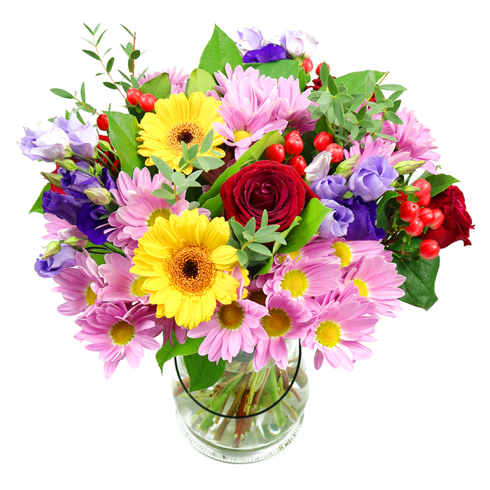 A lavish mixed bouquet stands in a clear glass vase, its colours radiating against a pristine white background like a pocket of spring in the middle of Streatham. Deep red roses nestle at the core, their velvety petals folded in intricate spirals that hint at long-held affection and meaningful moments. Just off-centre, bold yellow gerbera daisies with dark brown-orange centres face outward, their flat, open blooms exuding a sunny, modern charm that instantly lifts the mood. Soft pink daisies or chrysanthemums cluster around them, each flower made up of countless fine petals and small yellow eyes, adding a cushion of gentle colour and a feathery texture that softens the strong reds and yellows. Laced gracefully throughout the composition are delicate stems of purple and lavender lisianthus, their bell-shaped blossoms slightly ruffled at the edges, introducing a cool, sophisticated note reminiscent of twilight over Streatham Green. Bright red hypericum berries dot the bouquet like tiny beads of lacquer, delivering an unexpected, tactile accent that feels playful yet chic. The flowers are cradled in layered greenery, from broad deep-green leaves that frame the arrangement to lighter, more slender foliage that adds airiness and movement, as if caught in a soft breeze drifting across The Rookery gardens. The vase itself is simple and transparent, showing the cleanly cut stems submerged in clear water, evidence of a florist's meticulous preparation. This arrangement feels both exuberant and thoughtful, suited to celebrations, heartfelt thank-yous, or simply bringing a burst of colour and life into a Streatham home or office, where it can sit proudly on a table and quietly transform the room.